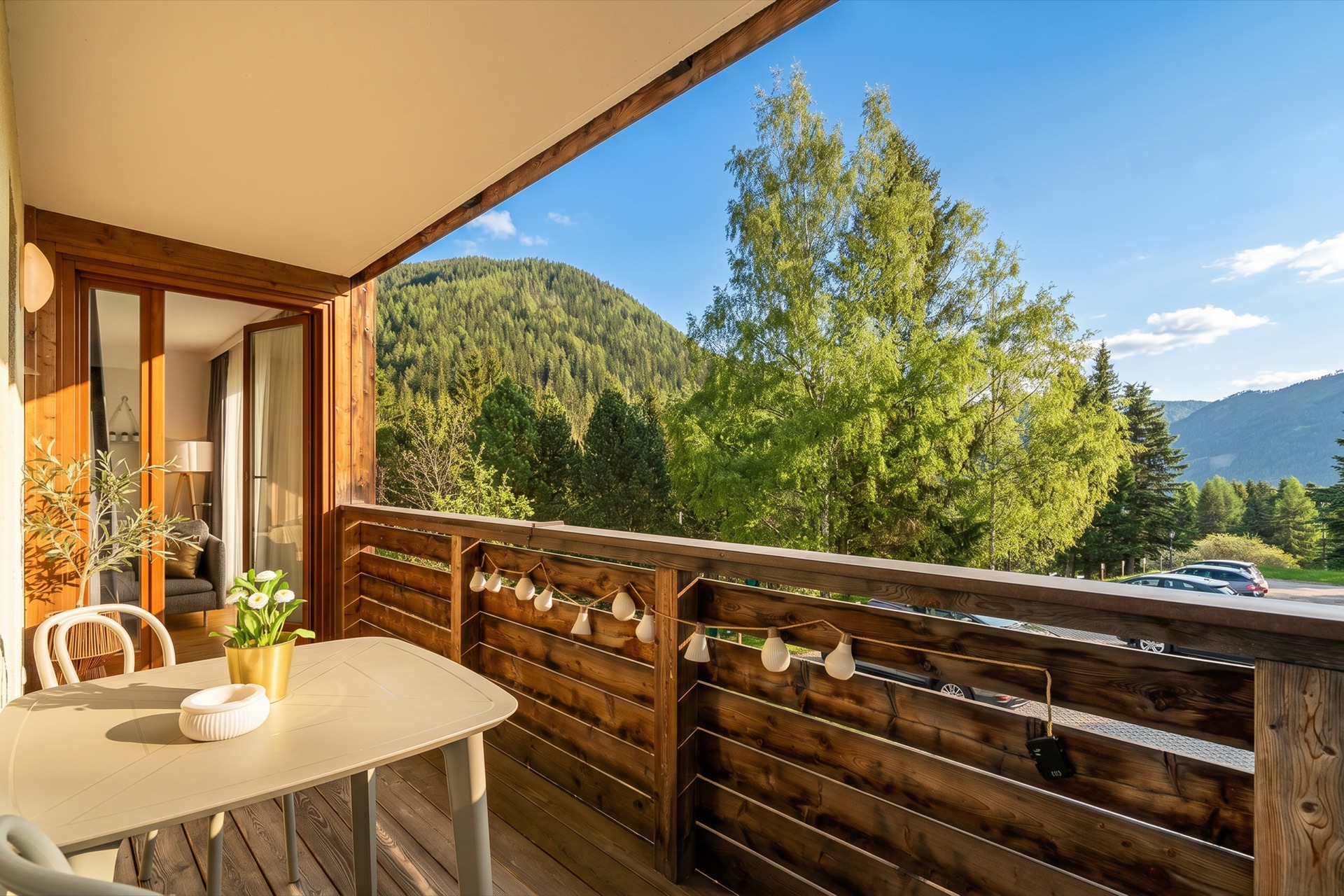 Holzbalkon mit weißem Bistrotisch, Lichterkette und warmem Abendlicht, Blick in das gemütliche Wohnzimmer und auf grüne Berglandschaft beim Alpstay Chalet Nr.1 Bad Kleinkirchheim – Balkonfotografie Ferienwohnung