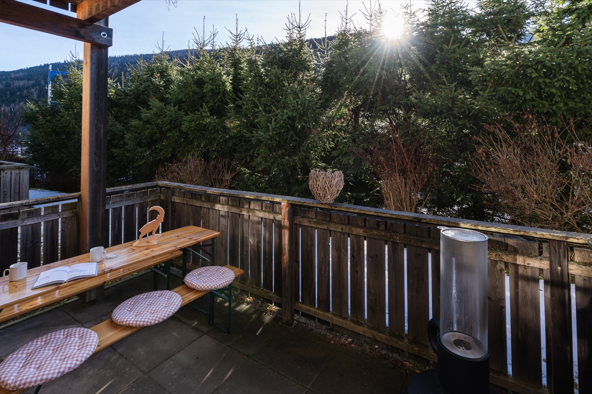 Sonnige Holzterrasse mit Bierzeltgarnitur, Sitzkissen und strahlendem Sonnenstern zwischen alpinen Bäumen der Ferienwohnung Holländer Kärnten – Terrassenfotografie Ferienwohnung